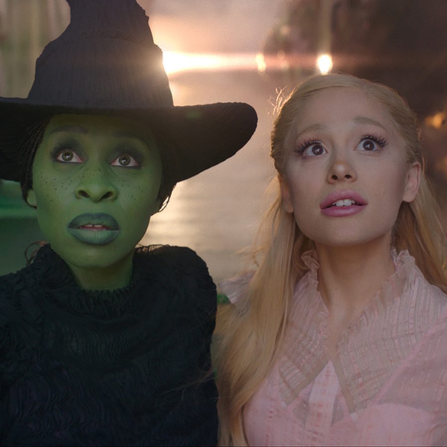  A scene from Wicked shows main characters Elphaba (Cynthia Erivo), a green-skinned witch dressed in black, and Galinda (Ariana Grande) a blonde witch dressed in pink, entering a grand room.  